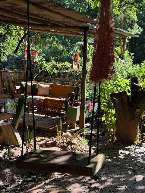 Balanço de madeira sob um abrigo rústico com assentos e vegetação exuberante.