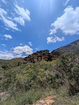 Afloramento rochoso em uma encosta sob um céu azul brilhante com nuvens dispersas. Folhagem verde em primeiro plano.