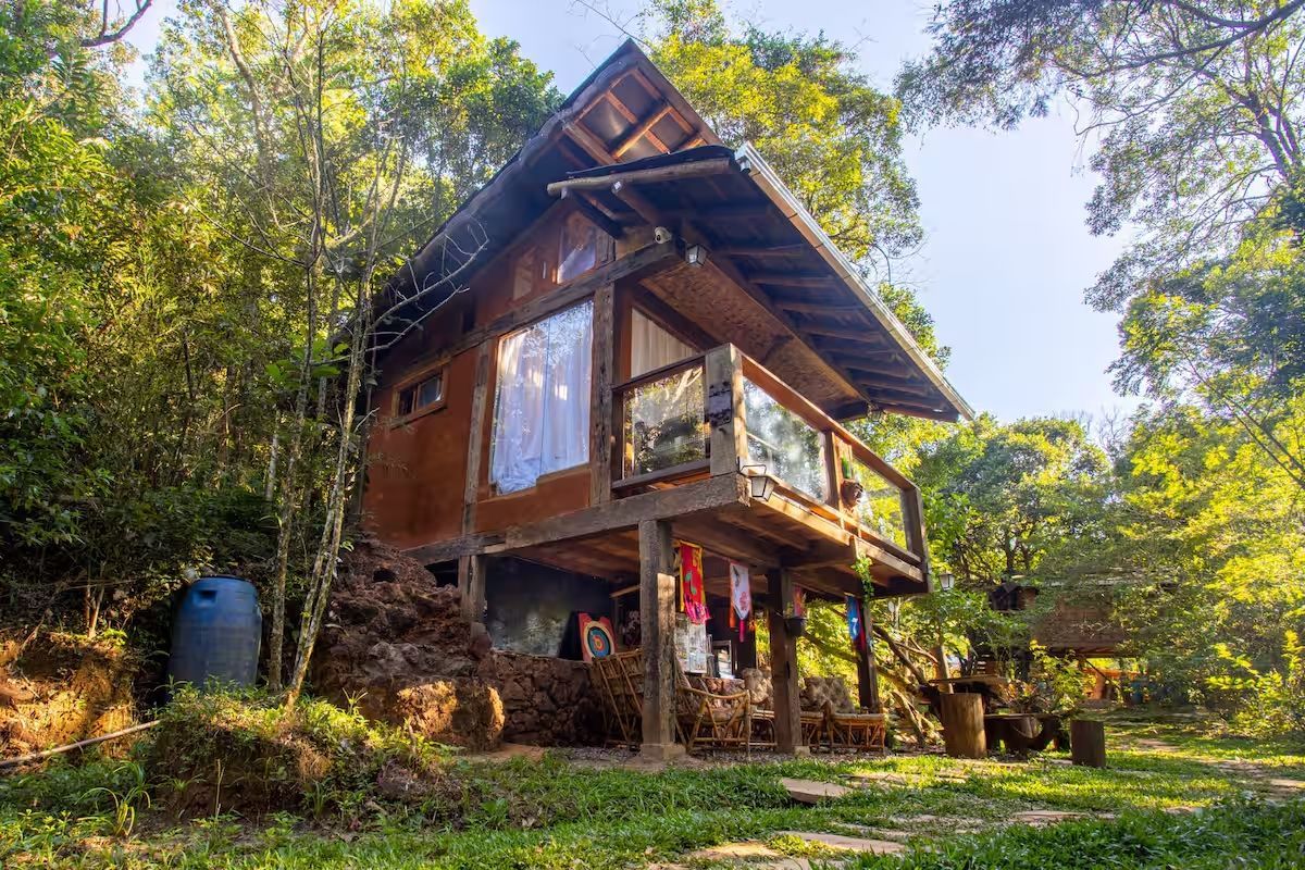 Cabana de madeira em meio à floresta, com varanda. A luz do sol penetra pelas frestas das árvores.
