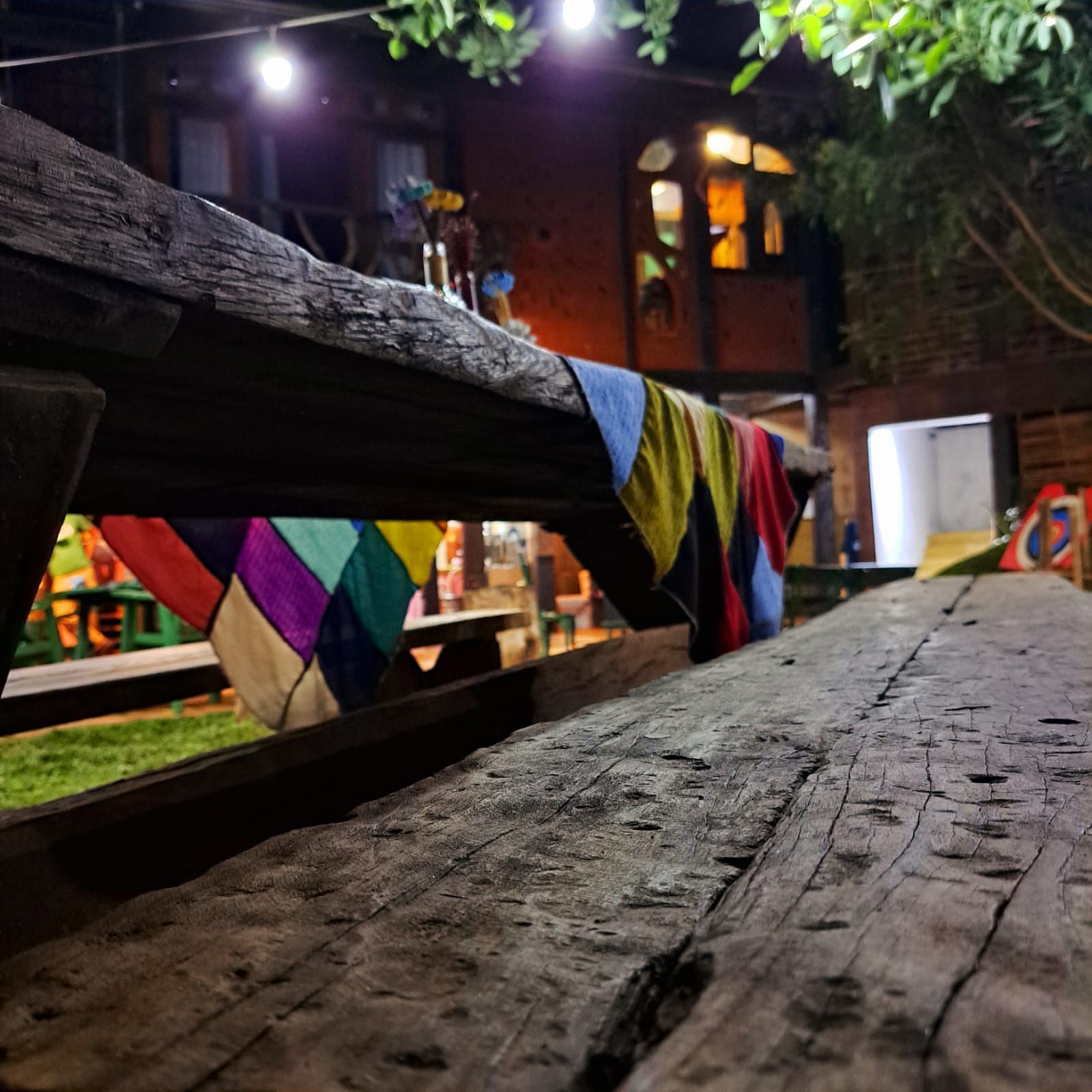 Mesa de piquenique de madeira em primeiro plano, vista do quintal à noite, tecido colorido pendurado e prédio com luzes.