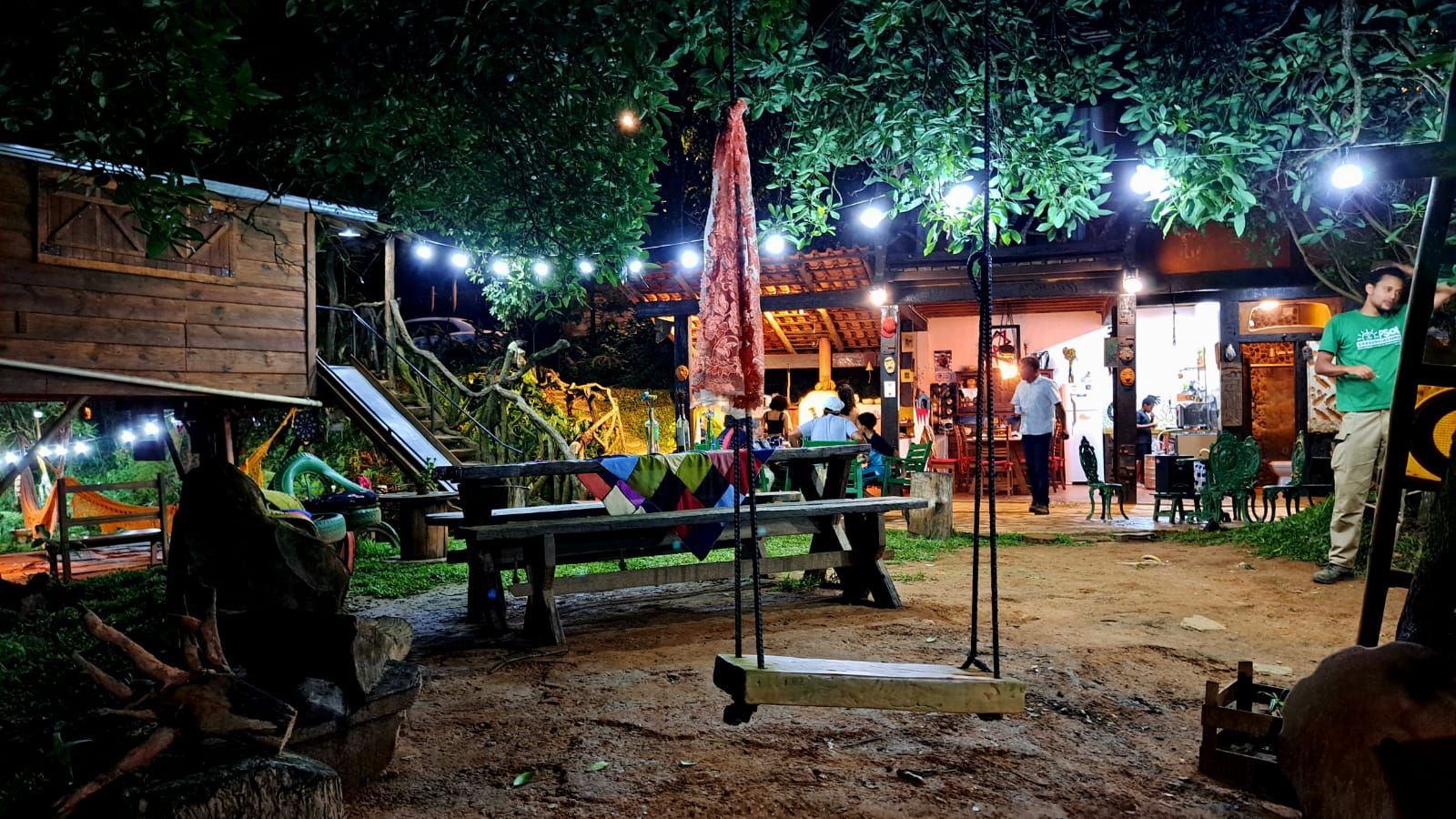 Restaurante ao ar livre à noite, com luzes de fada e um balanço. Pessoas sentadas às mesas; um ambiente rústico e descontraído.