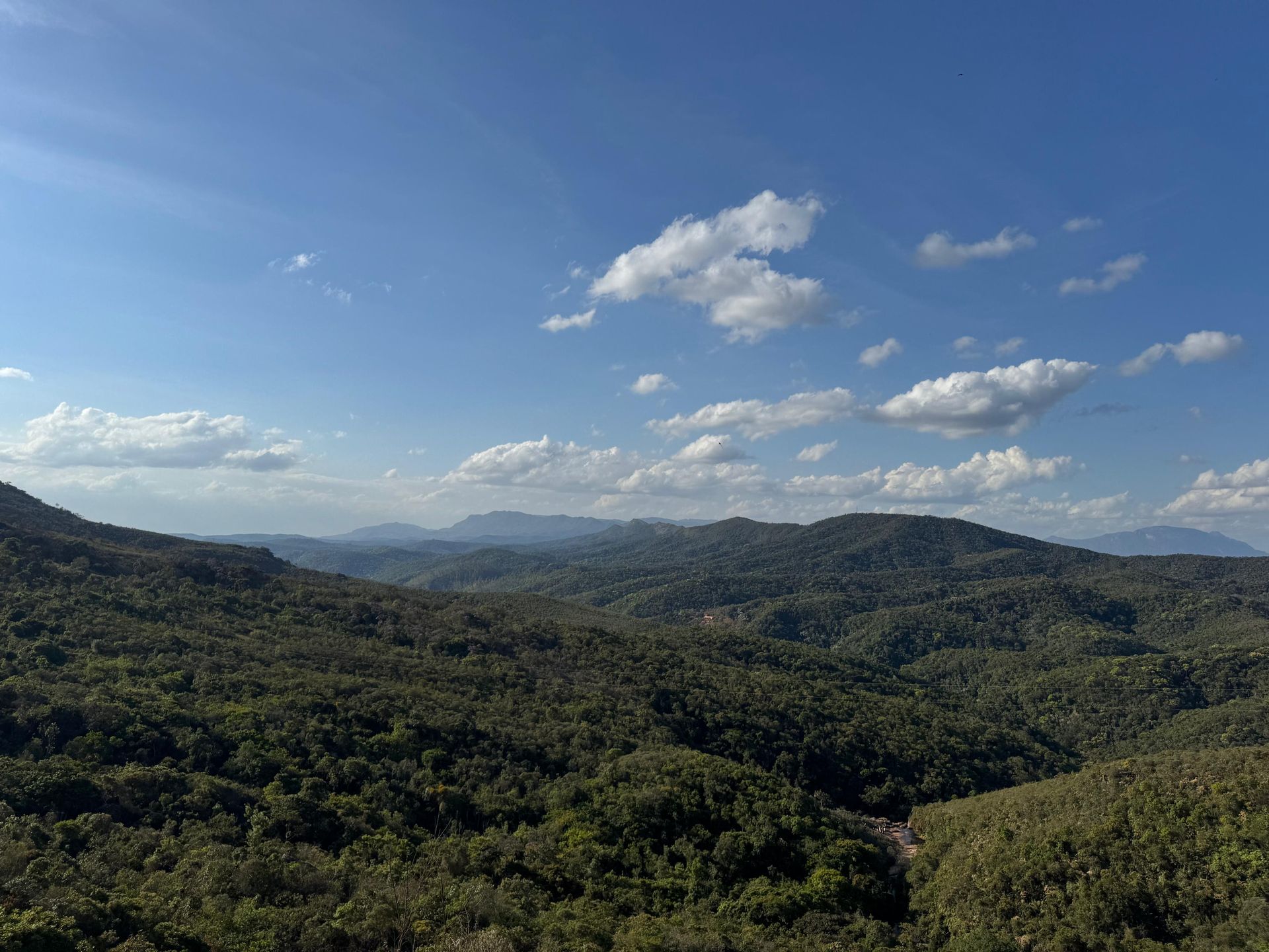 Vastas montanhas verdes cobertas de árvores sob um céu azul brilhante com nuvens brancas dispersas.