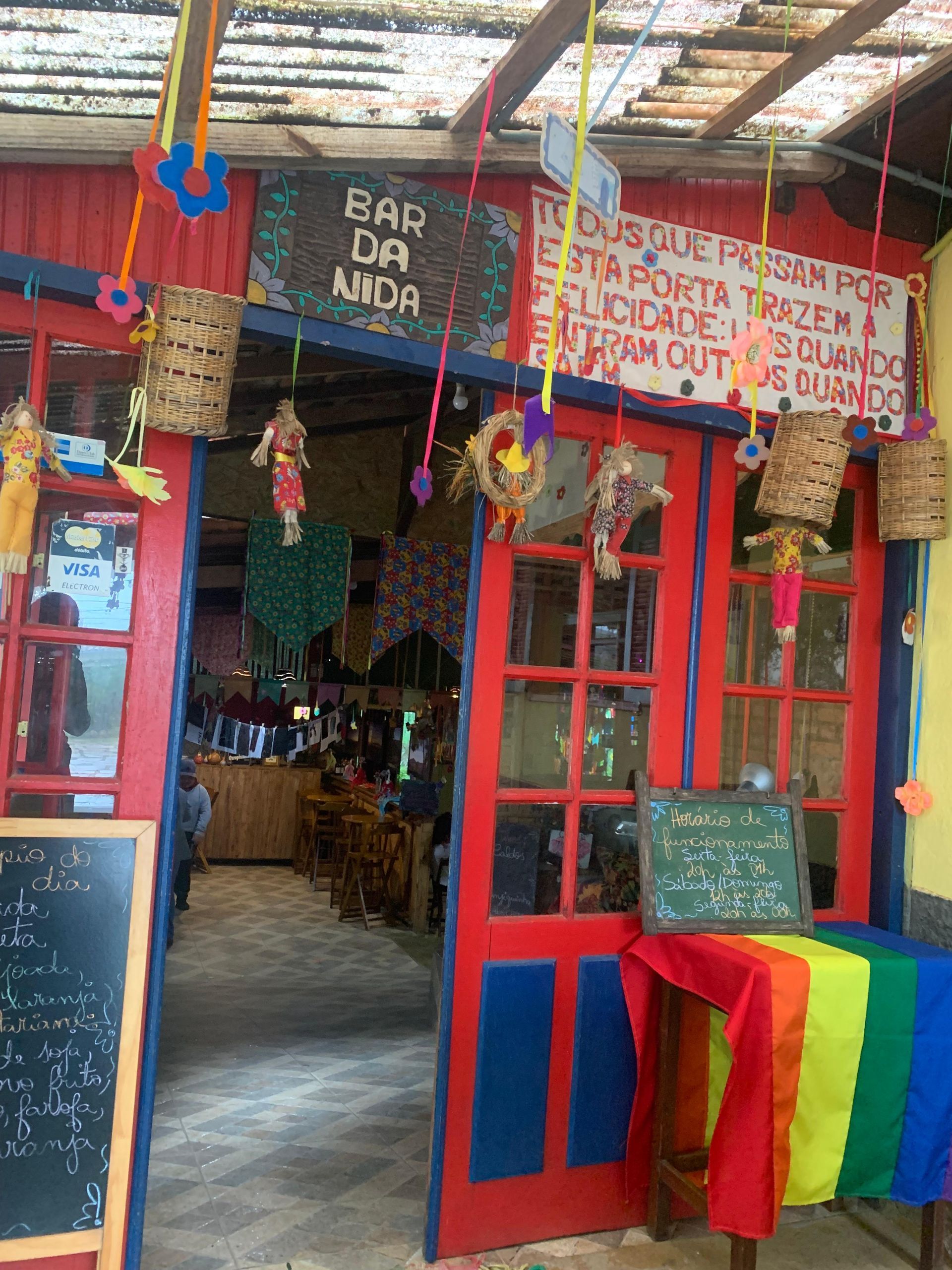 Entrada do bar pintada com cores vibrantes, portas vermelhas, mesa com as cores do arco-íris e decorações coloridas.