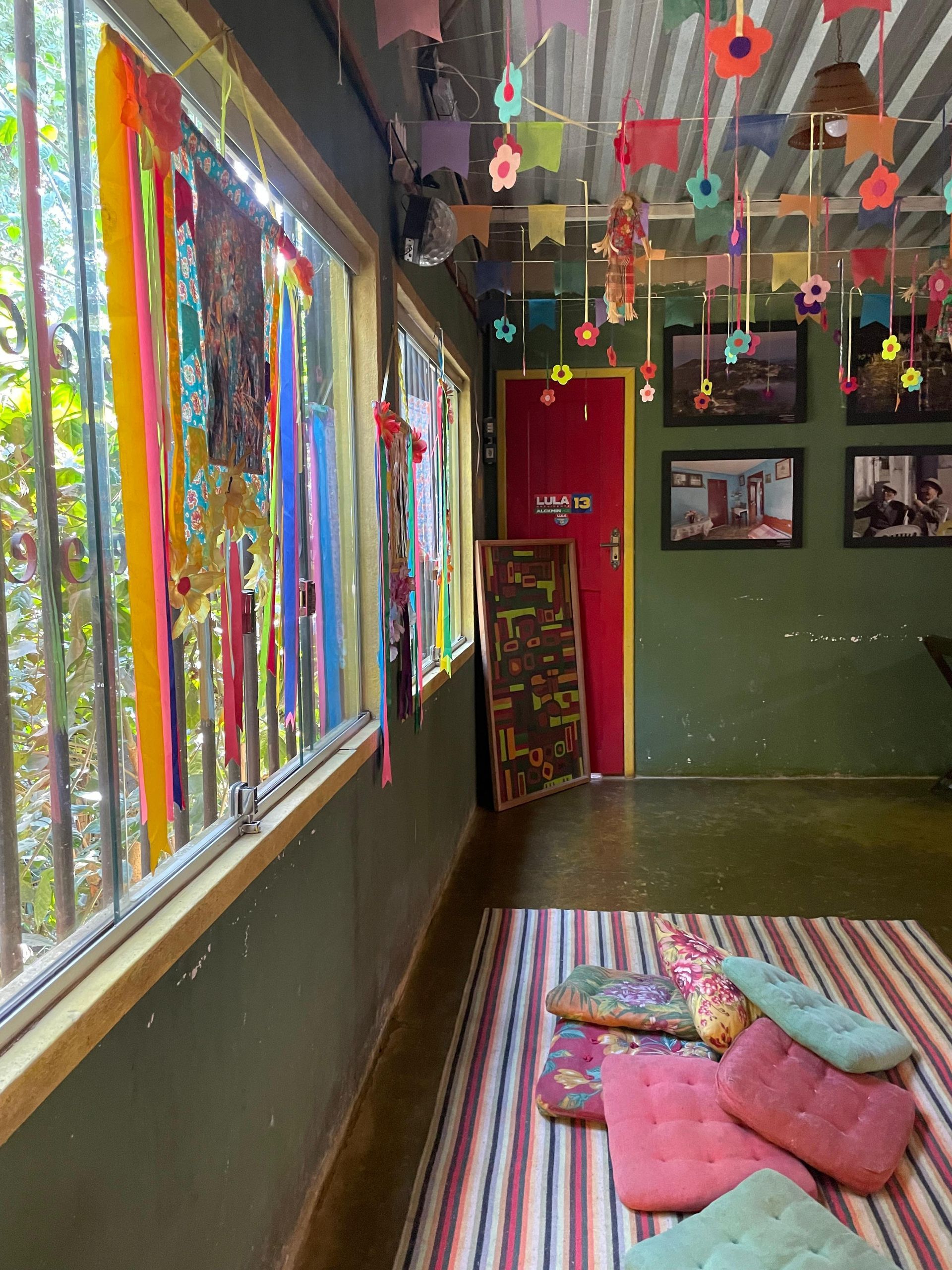 Quarto colorido com janela, decorado com flores de papel e tiras de tecido, com almofadas sobre um tapete listrado.