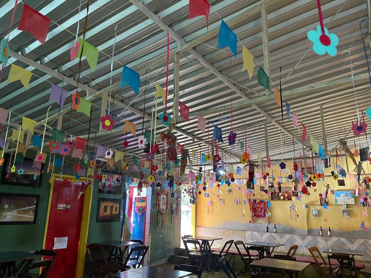 Interior decorado com bandeiras coloridas, flores e mesas; ambiente festivo, como de celebração.