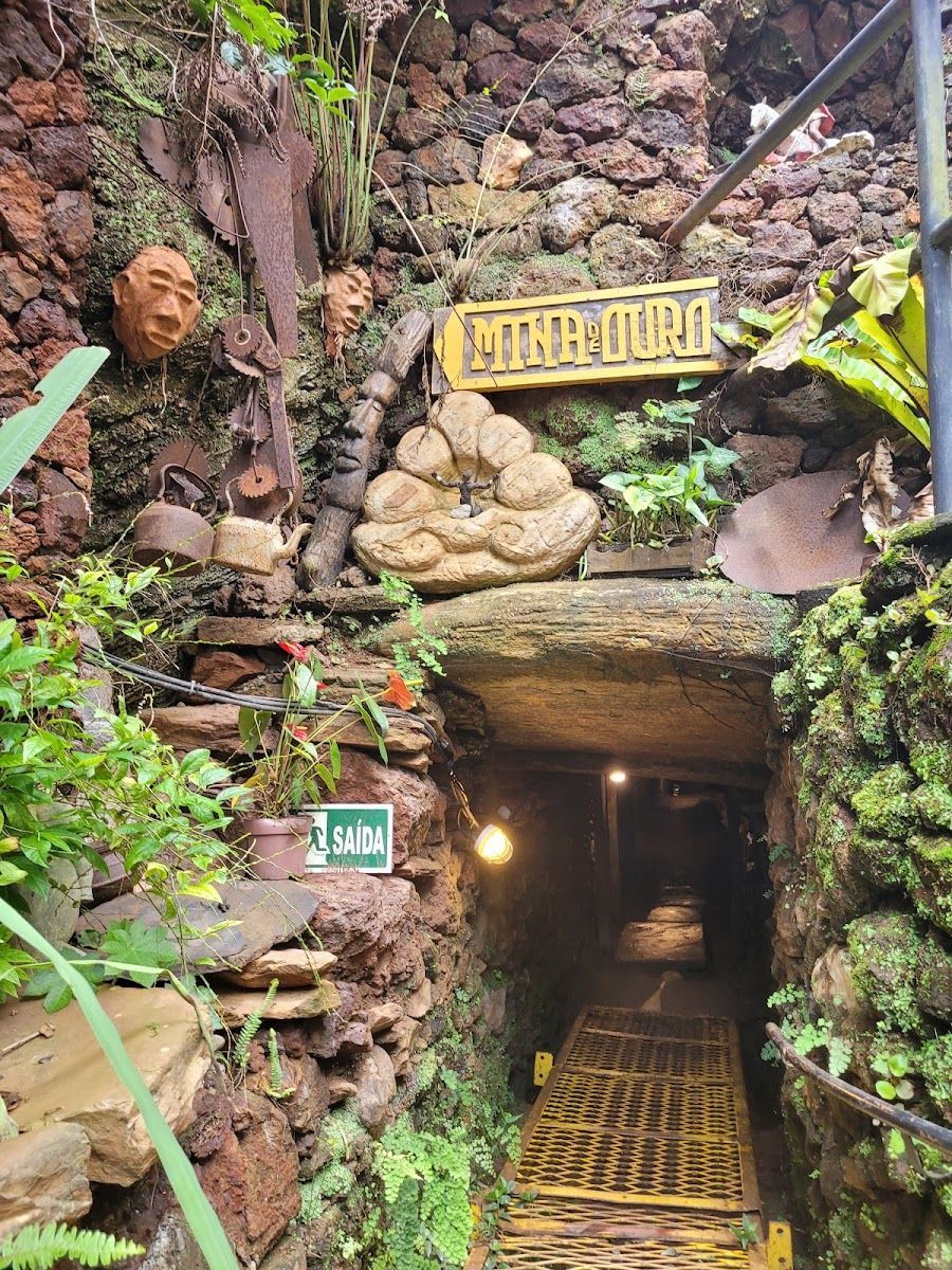 Entrada do túnel com paredes de pedra e vegetação. Placa de madeira e degraus conduzem ao interior.