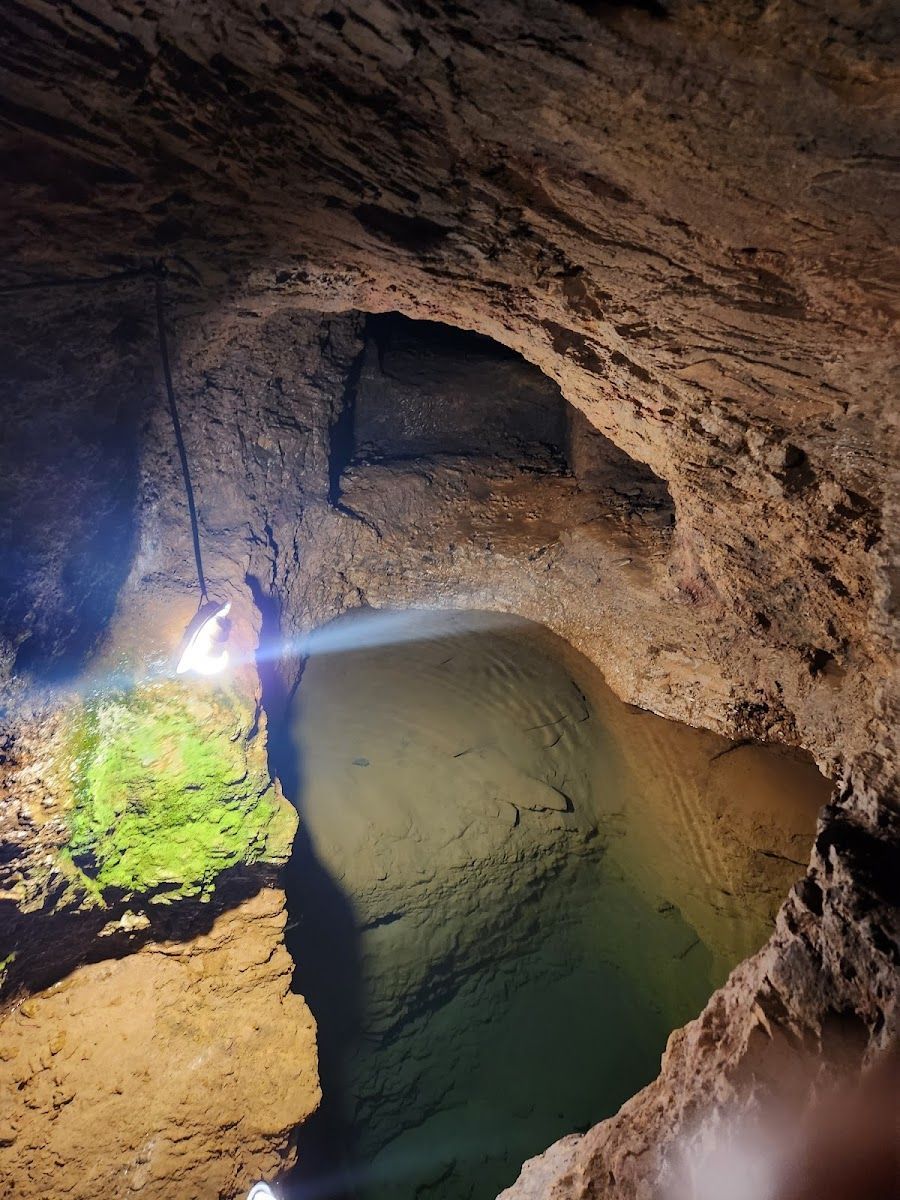 Caverna com um lago, onde a luz se reflete na superfície. Paredes rochosas e áreas cobertas de musgo.