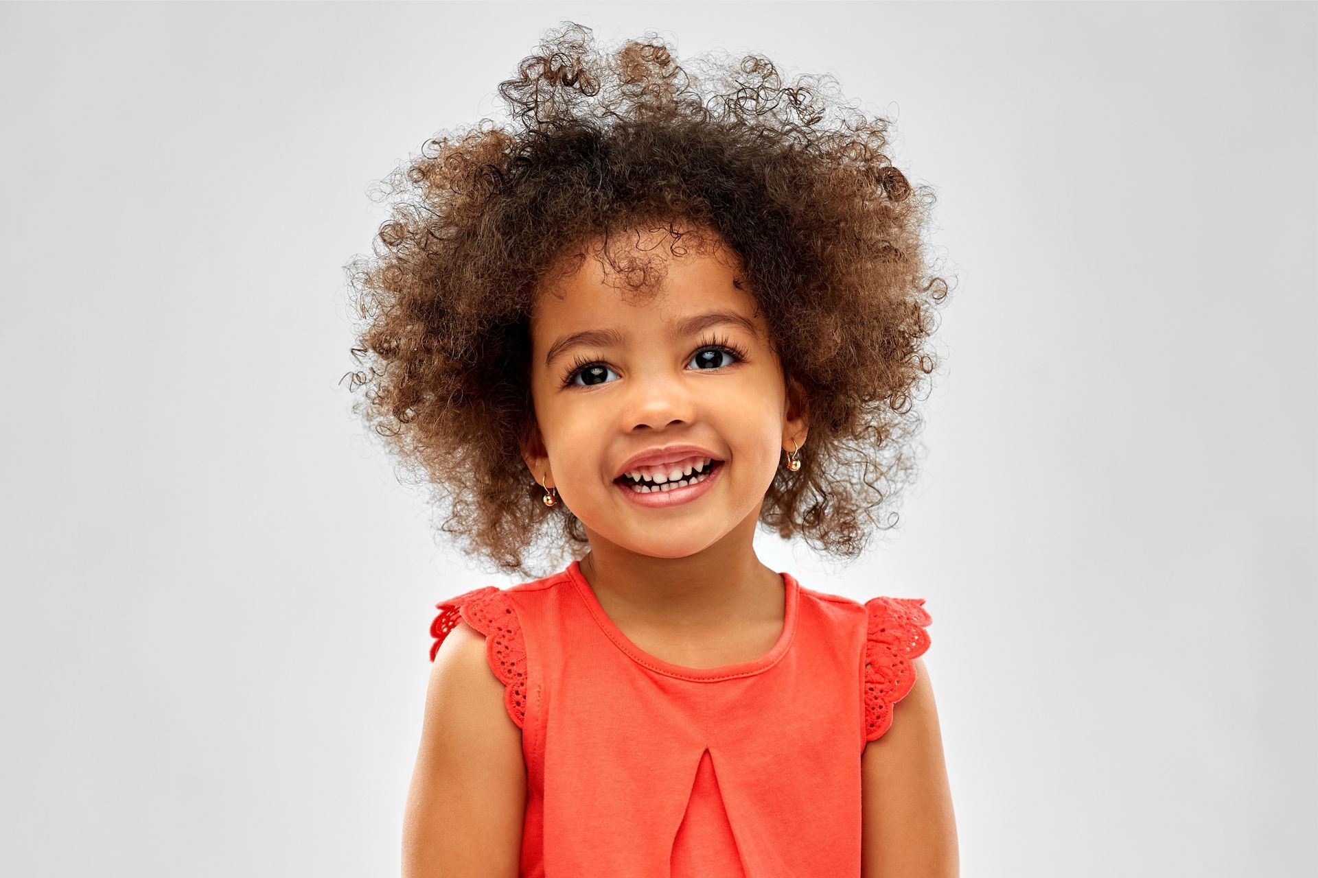 Smiling child with fluffy brown hair, wearing a red sleeveless top, against a light gray background. Smiling child with fluffy brown hair, wearing a red sleeveless top, against a light gray background.