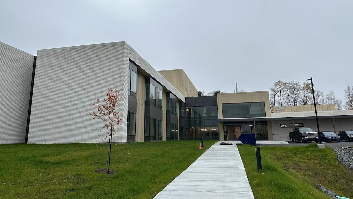 Vue extérieure d'un bâtiment moderne avec une façade en briques de couleur claire, des fenêtres en verre et un chemin en béton sur une colline herbeuse.