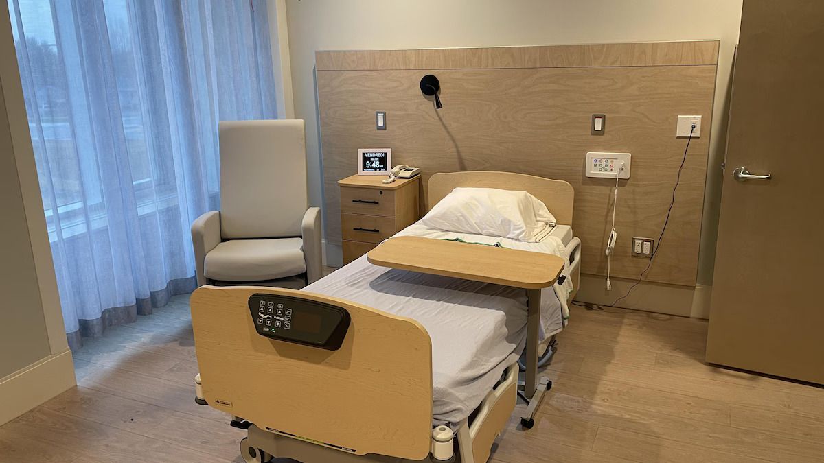 Chambre d'hôpital avec lit, chaise, table de chevet et fenêtre avec rideaux transparents.