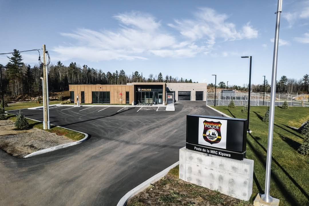 Un commissariat de police moderne avec un panneau devant. Une allée asphaltée mène au bâtiment, entouré d'arbres.