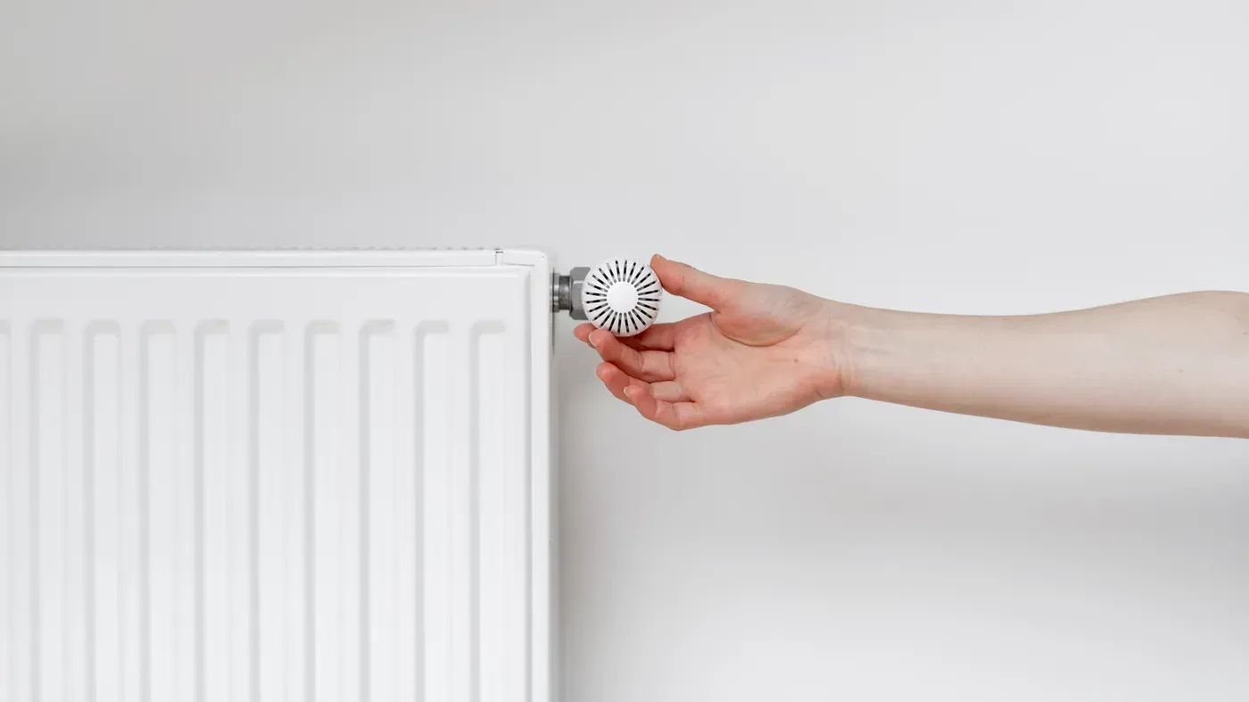 Une main actionne un thermostat sur un radiateur blanc.