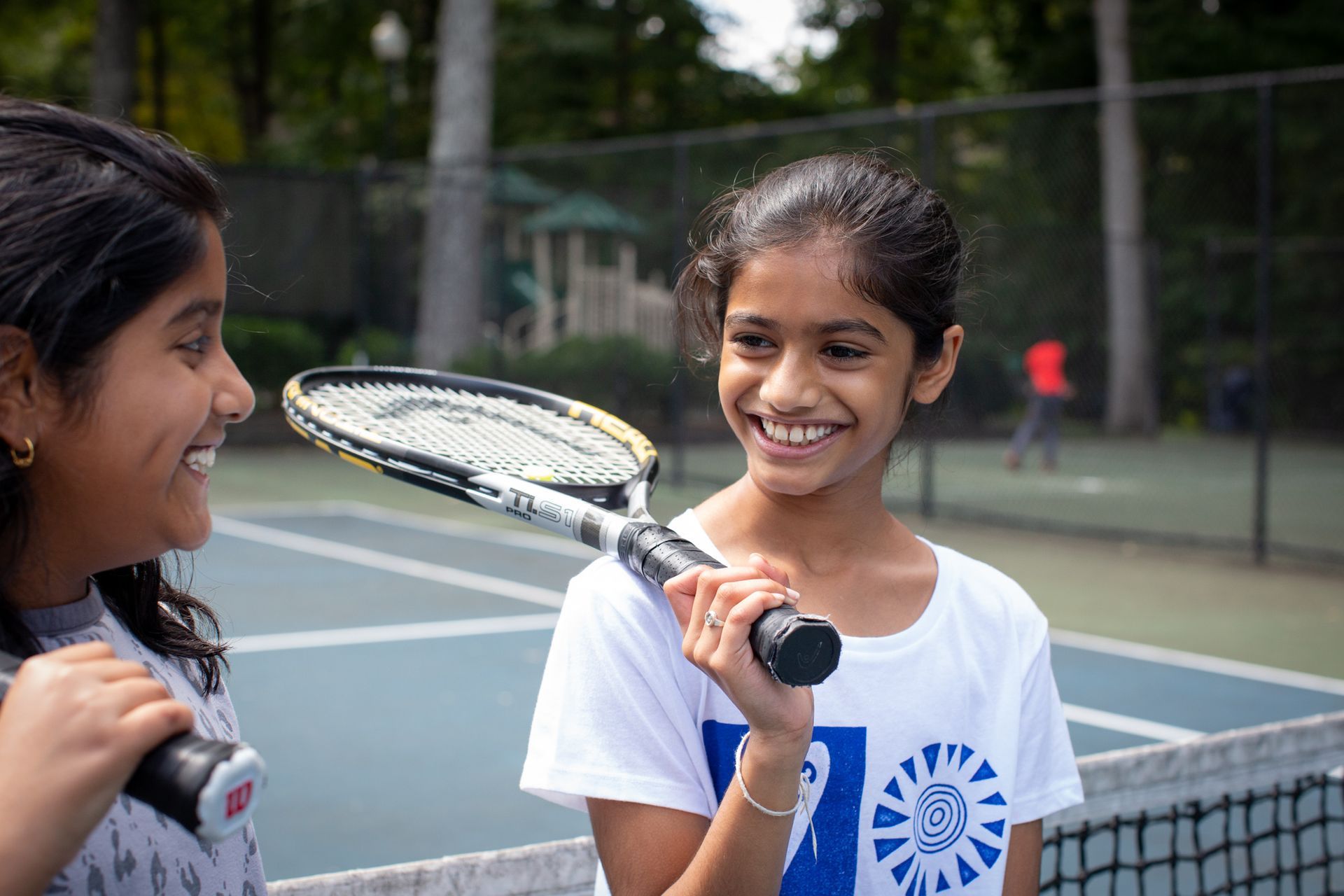 Two young girls are standing on a tennis court holding tennis rackets.