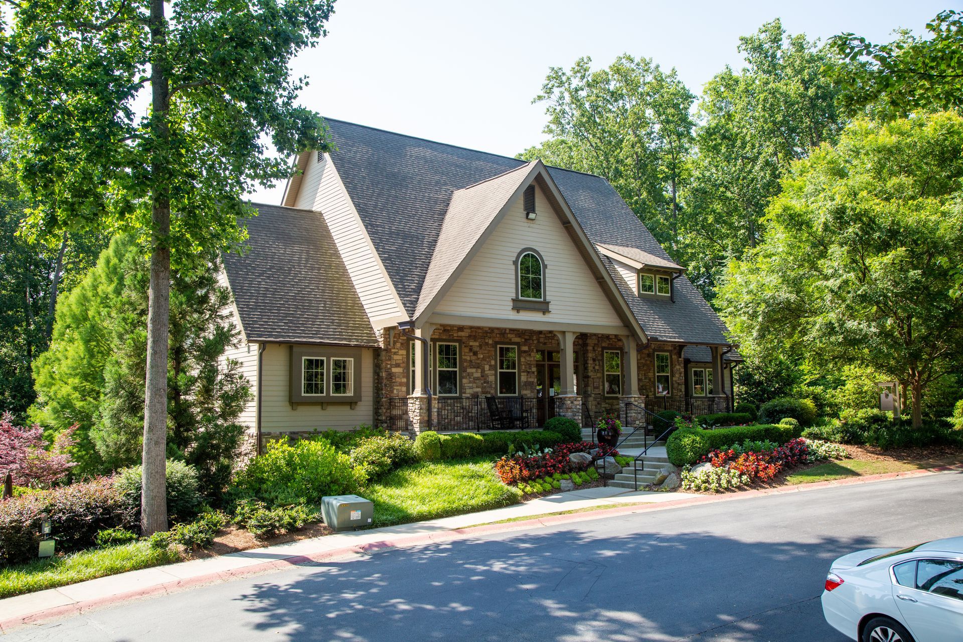 A white car is parked in front of a large house surrounded by trees.