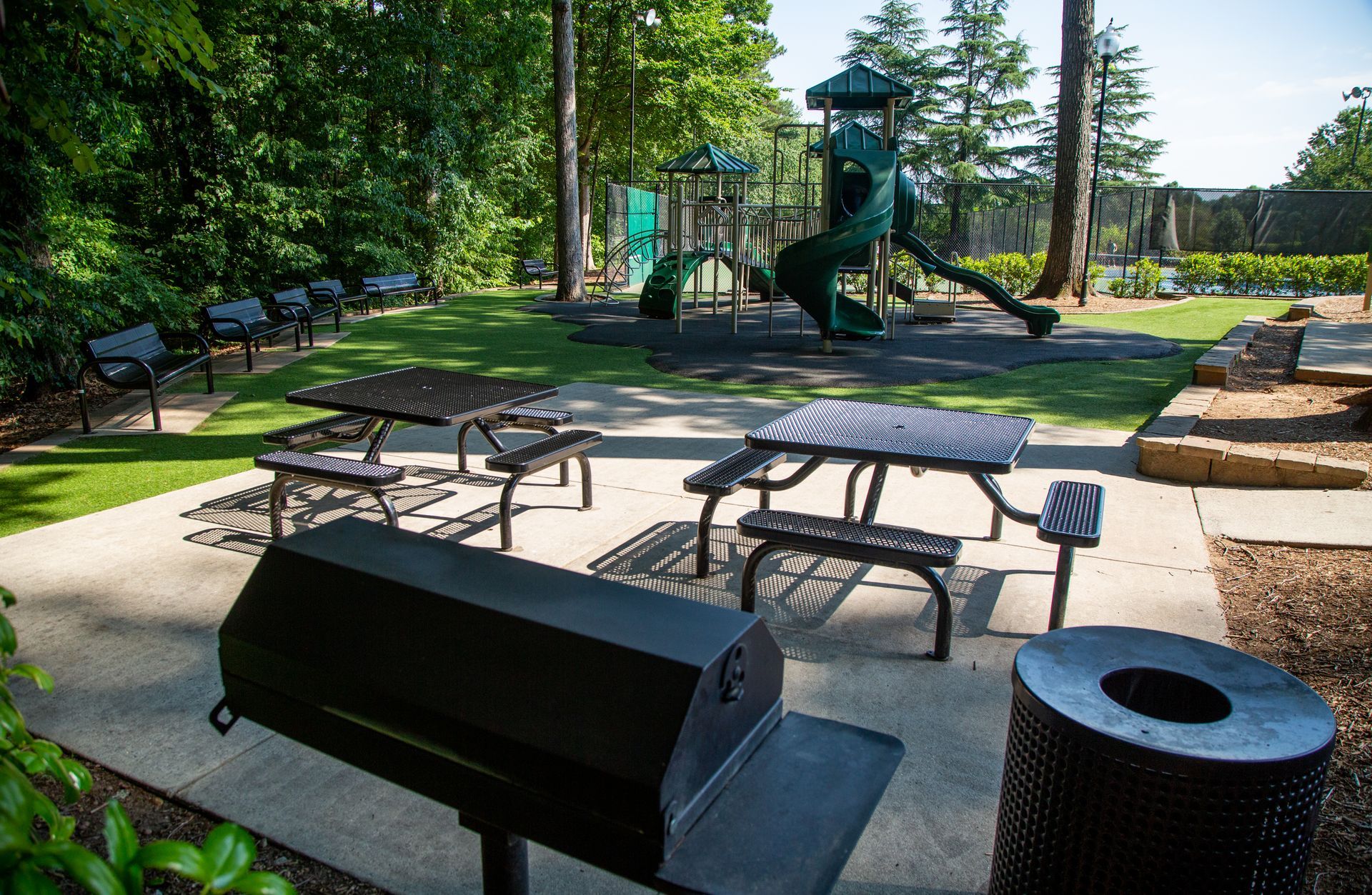 A picnic area with a grill and a trash can in front of a playground.