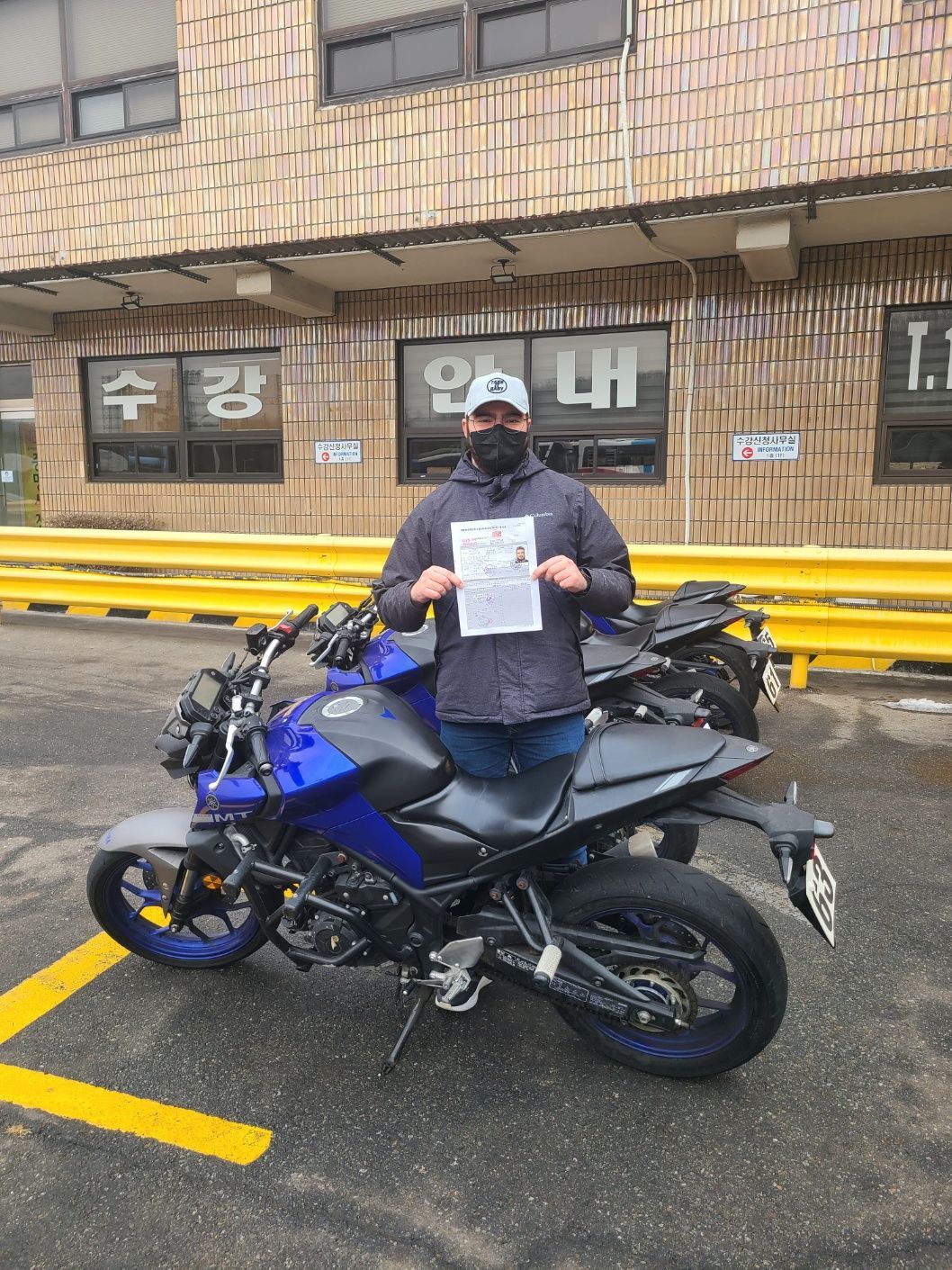 Person holding a paper, standing next to a blue motorcycle in front of a building with Korean writing.