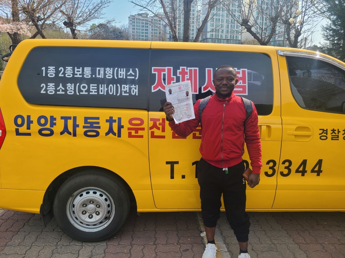 Man holding paper stands beside a yellow van. Van has Korean text and phone number.