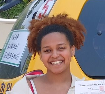 Woman smiling in front of a yellow vehicle with Korean text and a certificate.