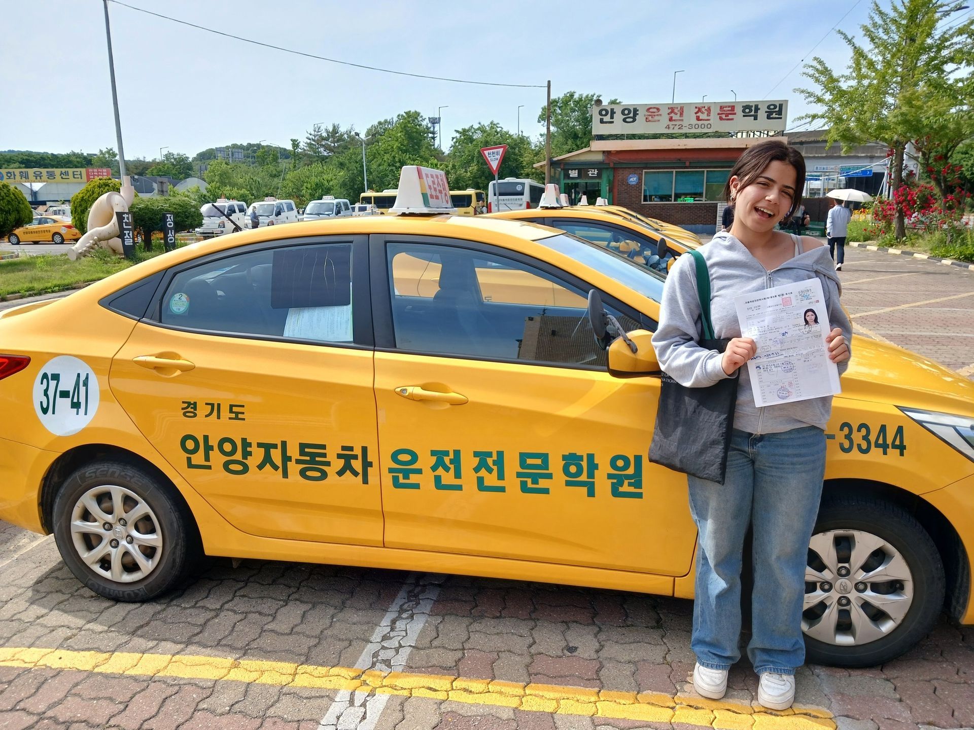 Woman holding a paper stands by a yellow taxi with Korean writing. Outside, sunny day.