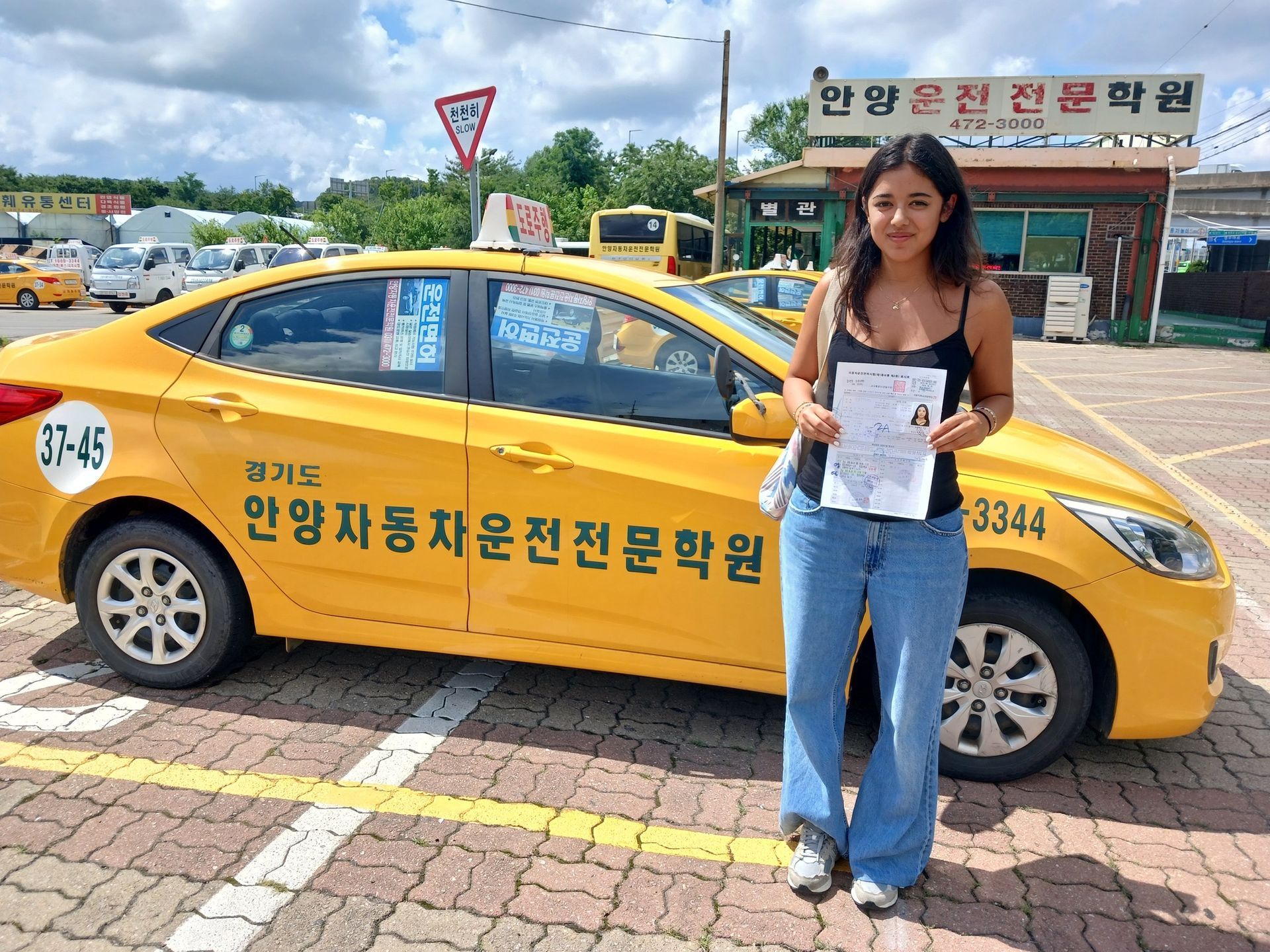 Woman holding a paper stands beside a yellow car, likely after a driving test, in a parking lot.