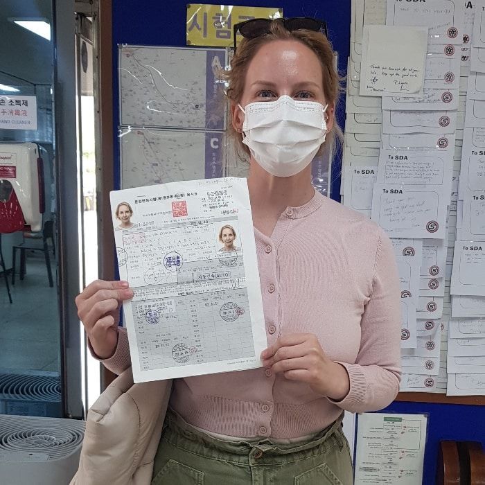 Woman in mask holding document, posing in a room with a bulletin board.