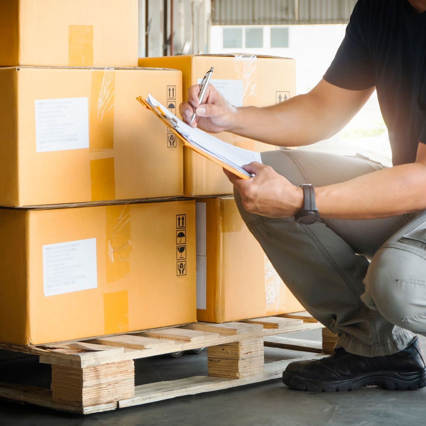 a man is kneeling down in front of a pallet of boxes and writing on a clipboard .