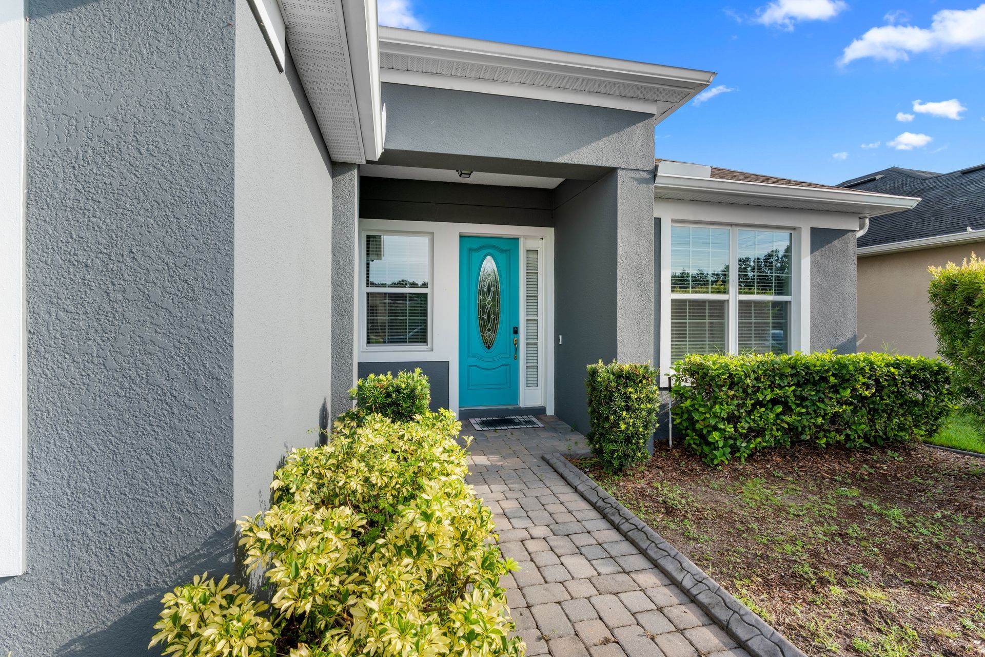 A teal front door on a gray house with a brick walkway, bushes, and windows.