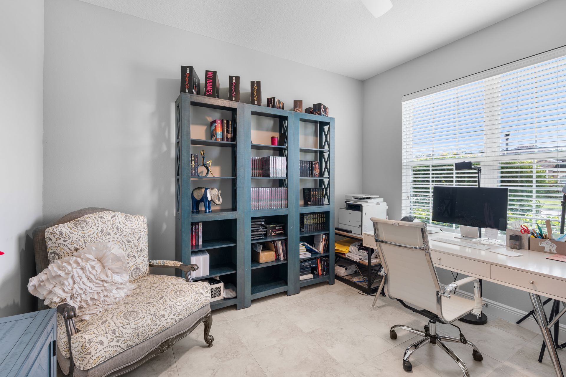 Home office with a large blue bookshelf, desk, chair, and patterned armchair by the window.