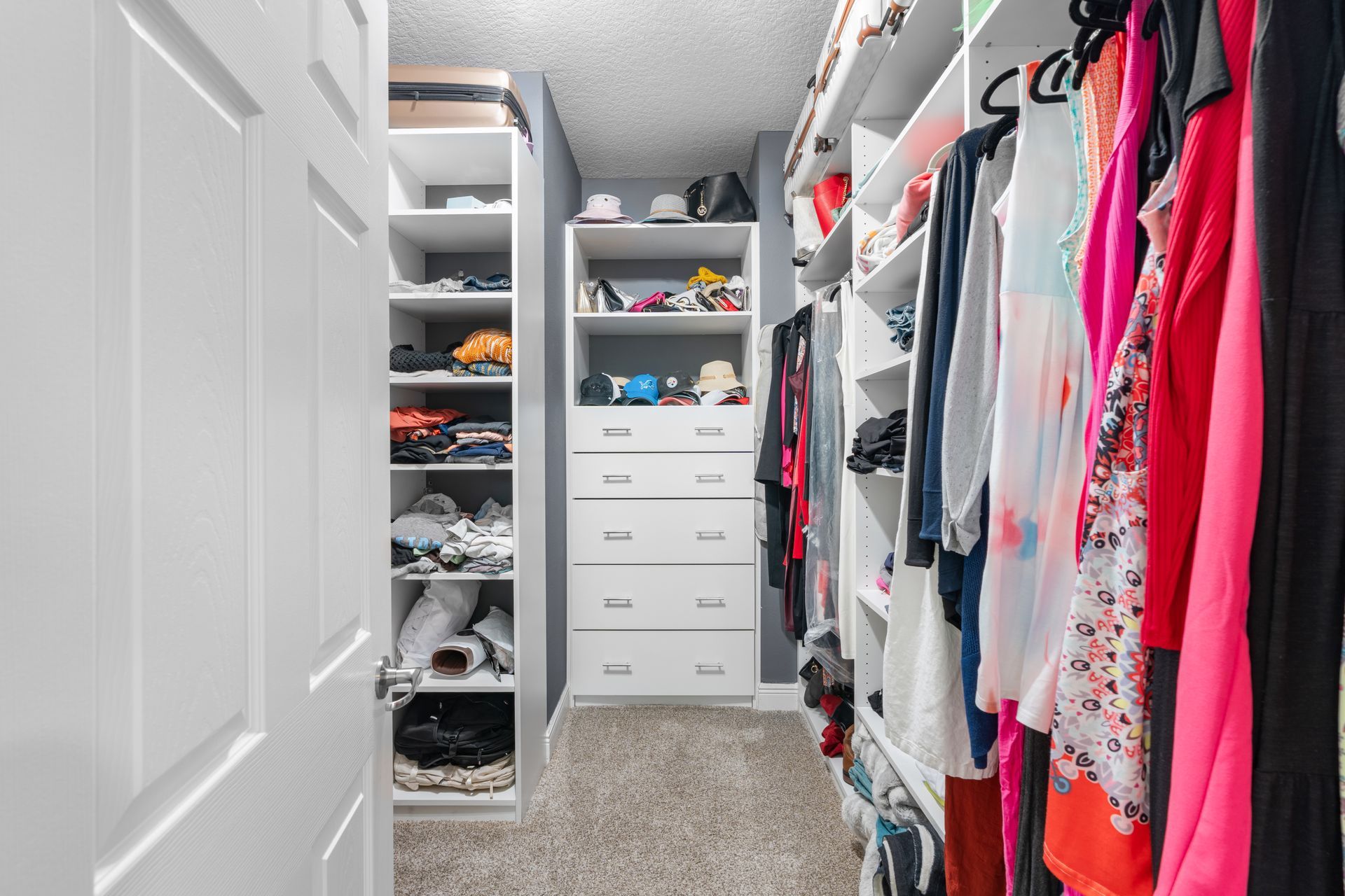 Walk-in closet with white shelving, clothing, and shoes. Grey walls, carpet, and ceiling.
