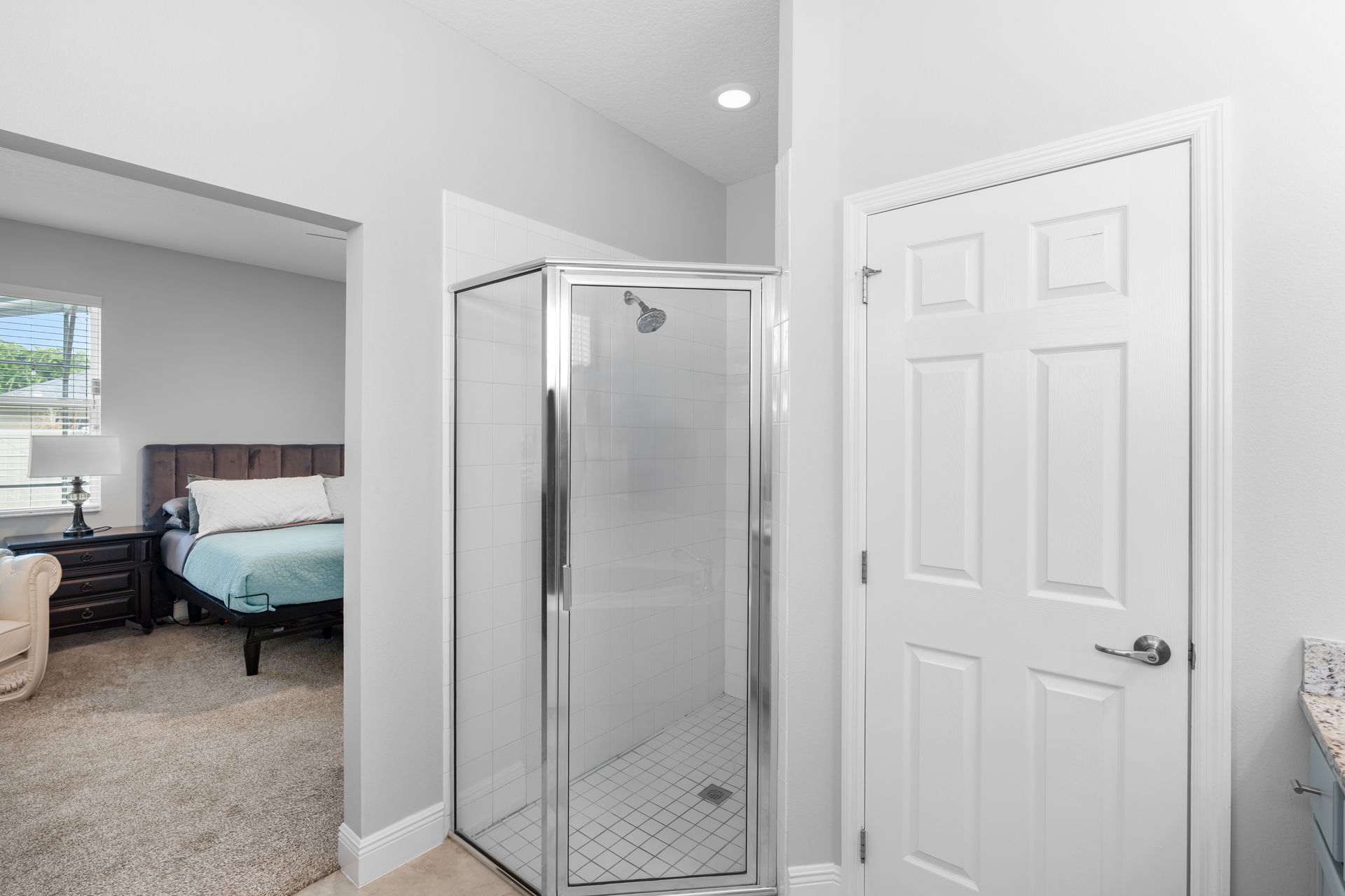Bathroom with a glass shower, white door, and view into a bedroom with a bed.