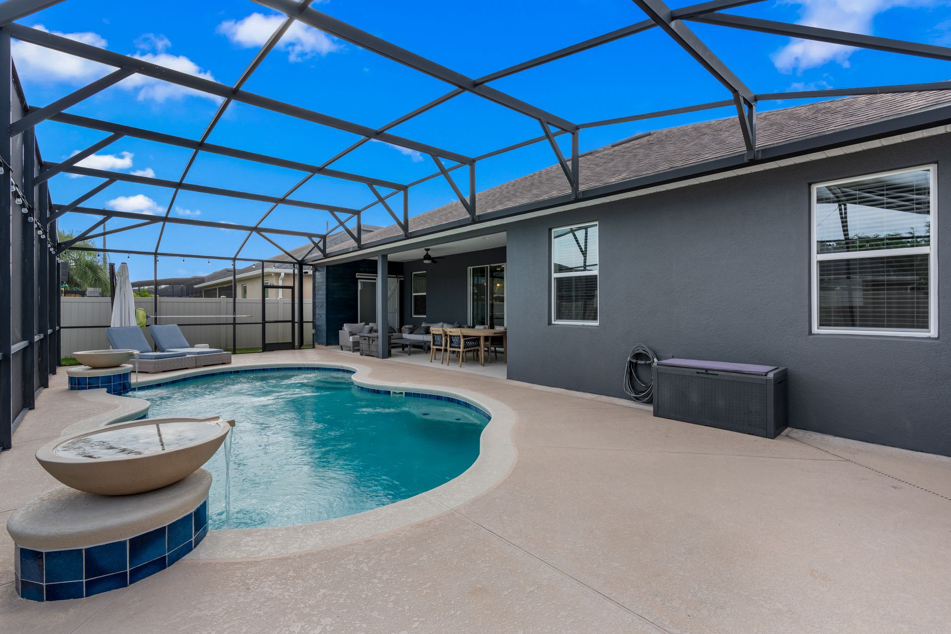 Pool area with blue water, screened enclosure, gray house, sunny day.