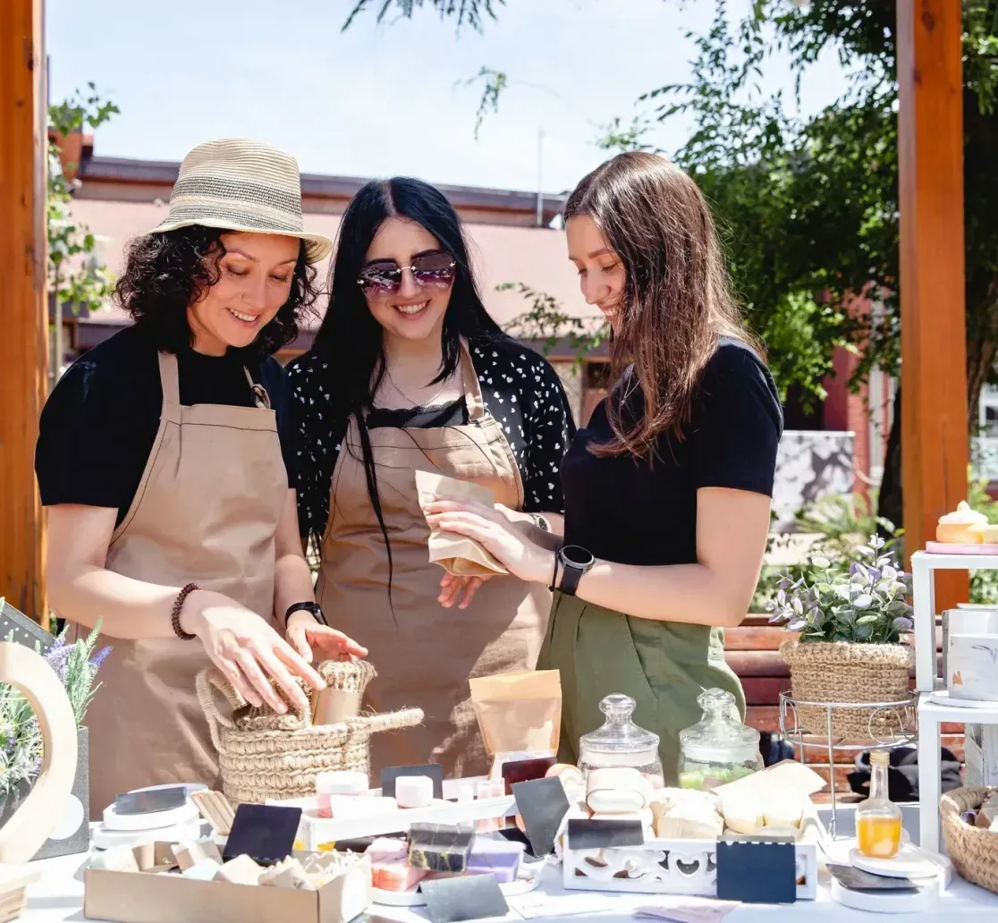 Three people in aprons stand behind a wooden stall outdoors, organizing and arranging artisanal soaps and products.