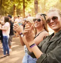 Two smiling people wearing sunglasses and festival wristbands clap while standing in a sunny, wooded outdoor venue.