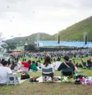 People sit on chairs and mats in a large outdoor field at a festival with a stage, mountains, and a ferris wheel nearby.