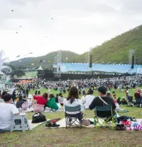 People sit on chairs and mats in a large outdoor field at a festival with a stage, mountains, and a ferris wheel nearby.