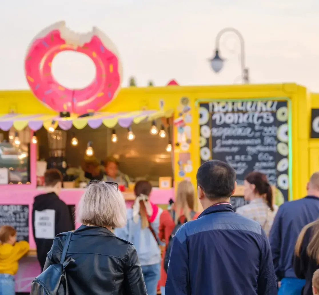 People stand in a line waiting at a bright yellow food truck decorated with a giant pink donut sign.