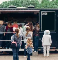 A crowd of people stands in line at the service window of a black food truck outdoors.