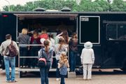 A crowd of people stands in line at the service window of a black food truck outdoors.