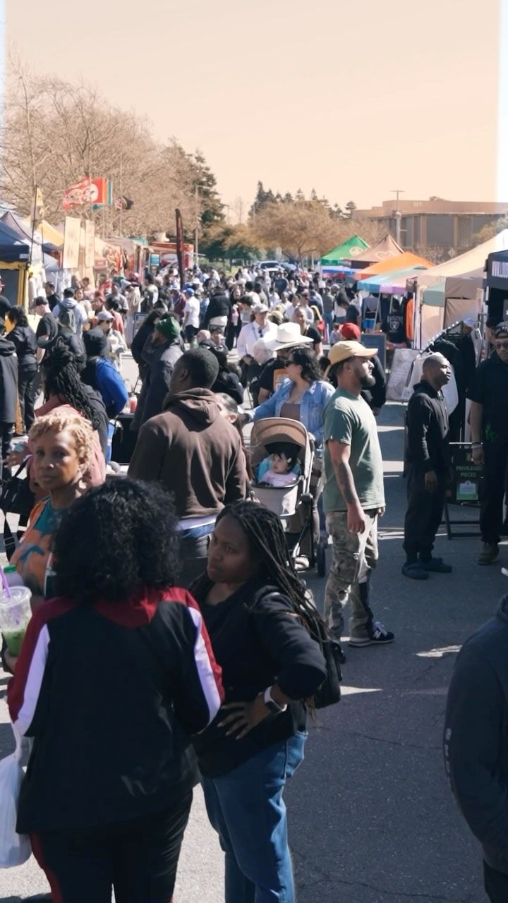 A crowded outdoor street market with people walking among vendor tents on a sunny day.