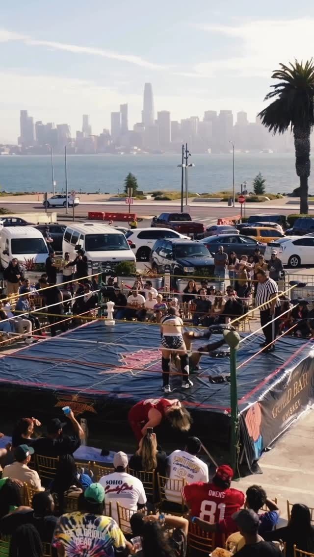A wrestling match takes place in an outdoor ring with the San Francisco skyline in the background.