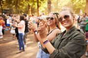Two smiling people wearing sunglasses and festival wristbands clap while standing in a sunny, wooded outdoor venue.