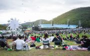 People sit on chairs and mats in a large outdoor field at a festival with a stage, mountains, and a ferris wheel nearby.
