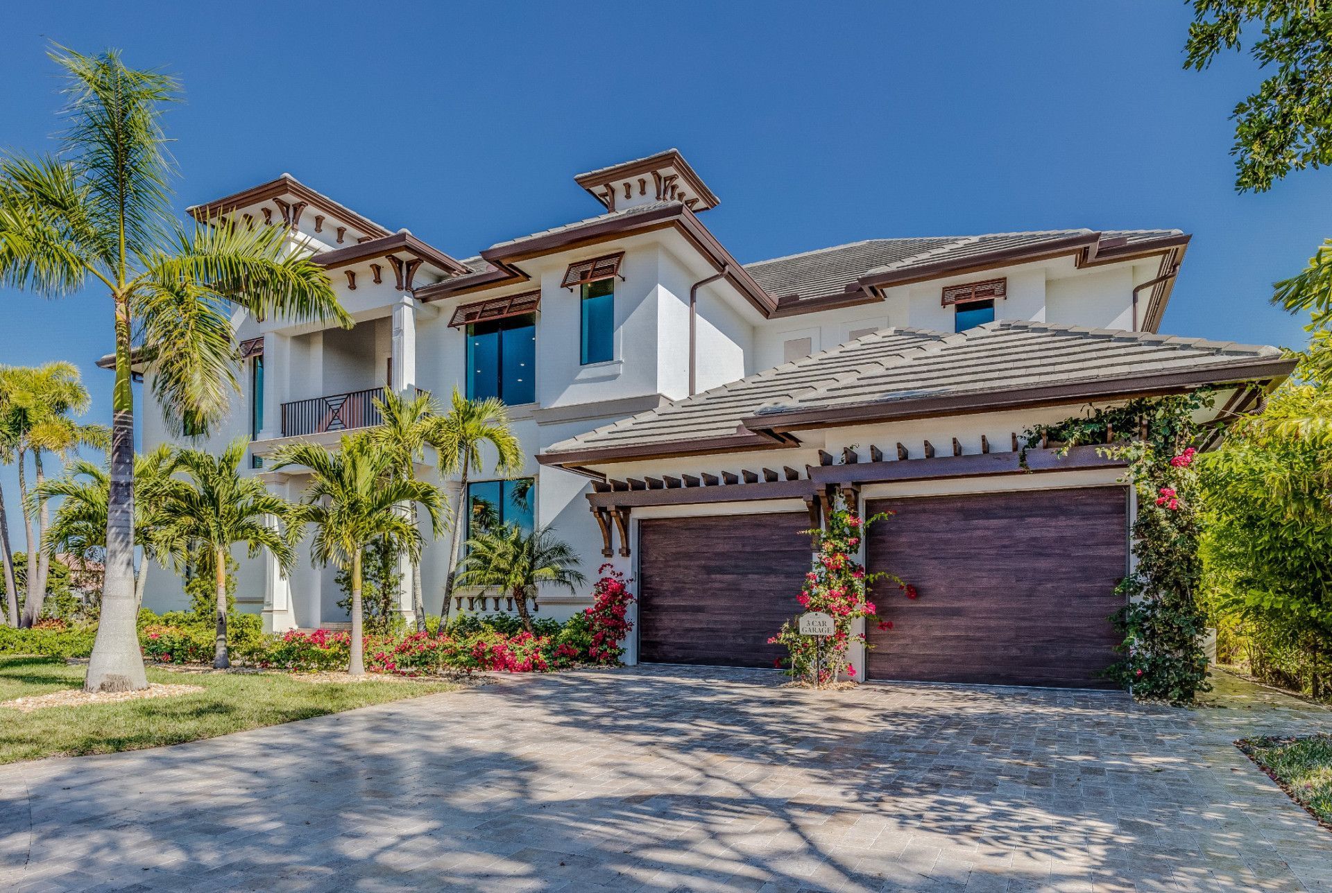 A large white house with two garage doors is surrounded by palm trees.