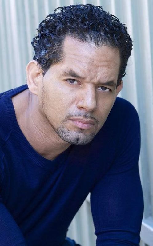 A man with curly hair and a beard is wearing a blue shirt and looking at the camera.