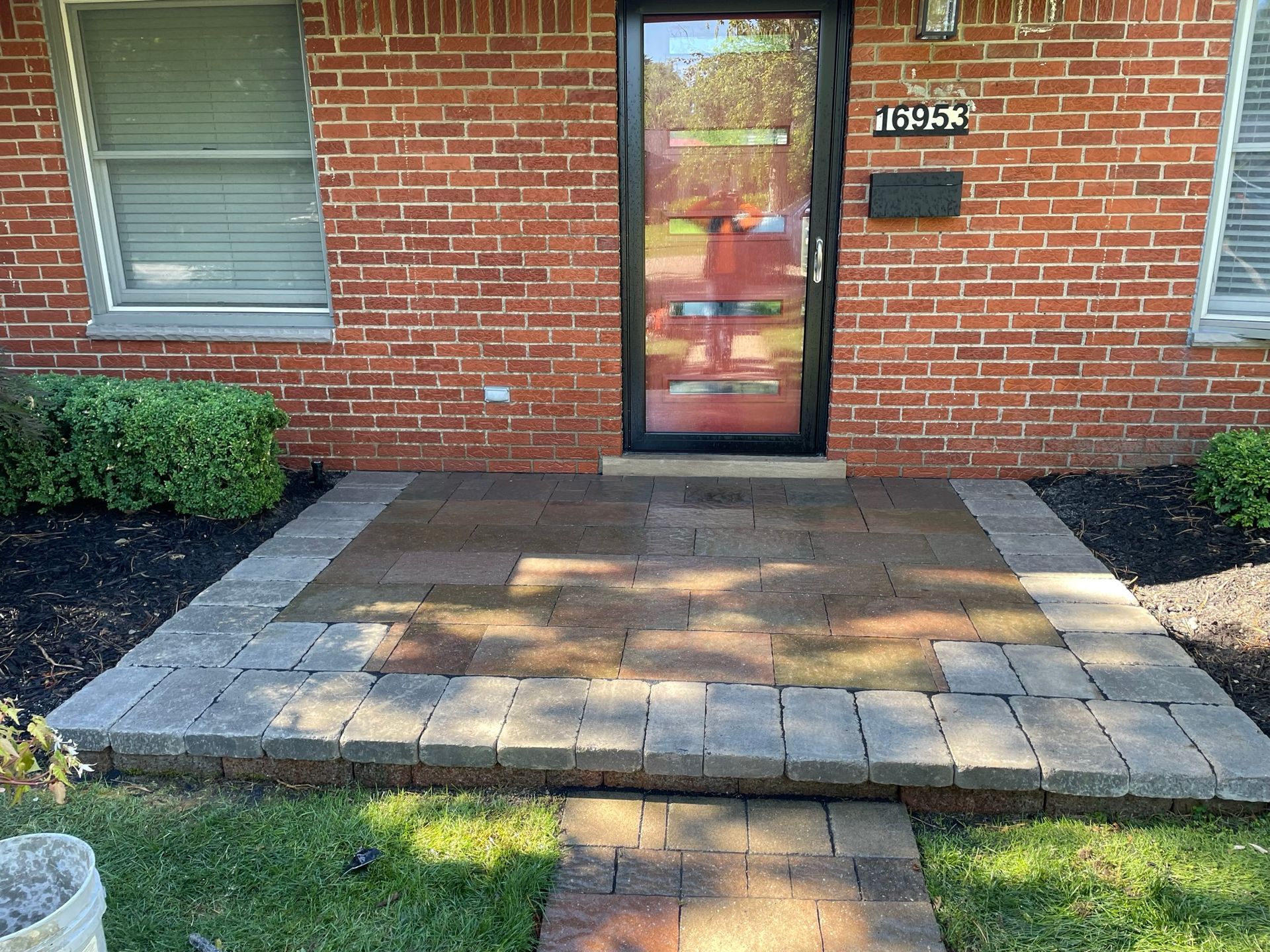 Brick home entrance with paved pathway and small steps. Dark door, address numbers, and garden beds with greenery.