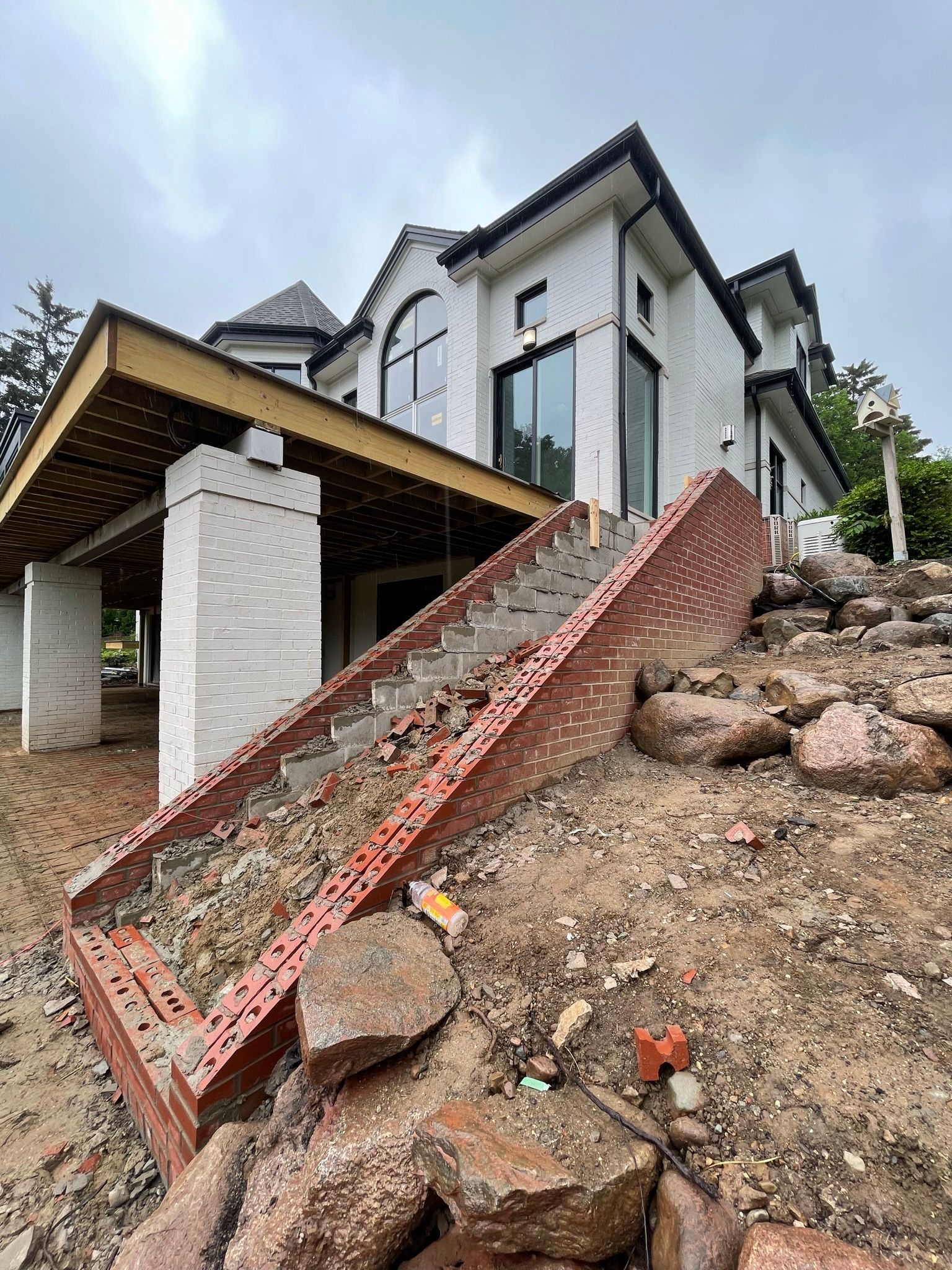 Brick stairs lead up to a modern white house under construction. The house sits on a hillside with exposed dirt and rocks.