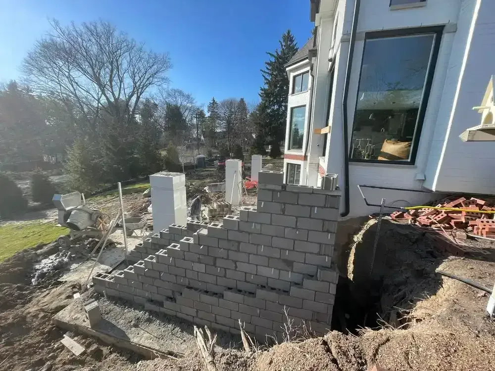 Construction site with a partially built brick retaining wall next to a white house; daylight and clear blue sky.