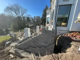 Construction site with a partially built brick retaining wall next to a white house; daylight and clear blue sky.