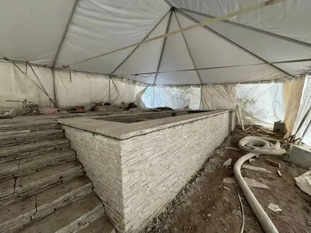A partially constructed pool with stone facade and steps, inside a temporary tent structure. Debris litters the ground.