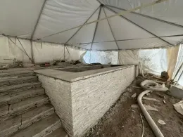 A partially constructed pool with stone facade and steps, inside a temporary tent structure. Debris litters the ground.